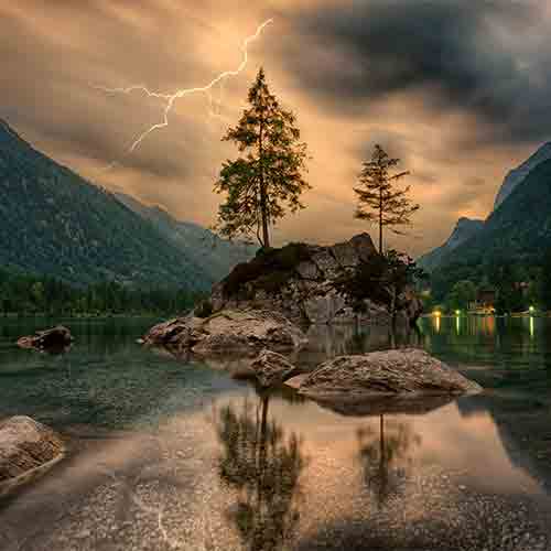 Lightning over a tranquil lake with two trees on a rocky islet at dusk